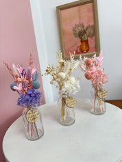 Three small glass vases with dried flowers on a white surface, with a framed picture in the background.