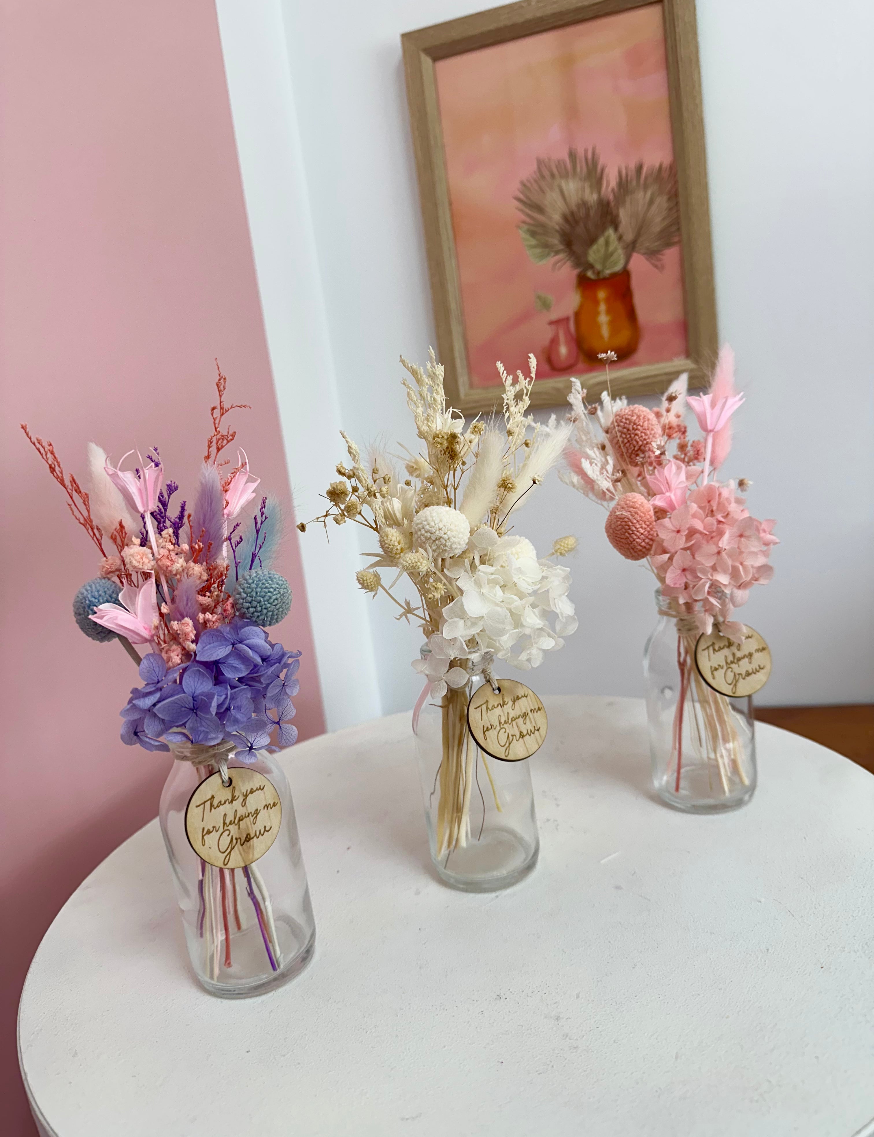Three small glass vases with dried flowers on a white surface, with a framed picture in the background.
