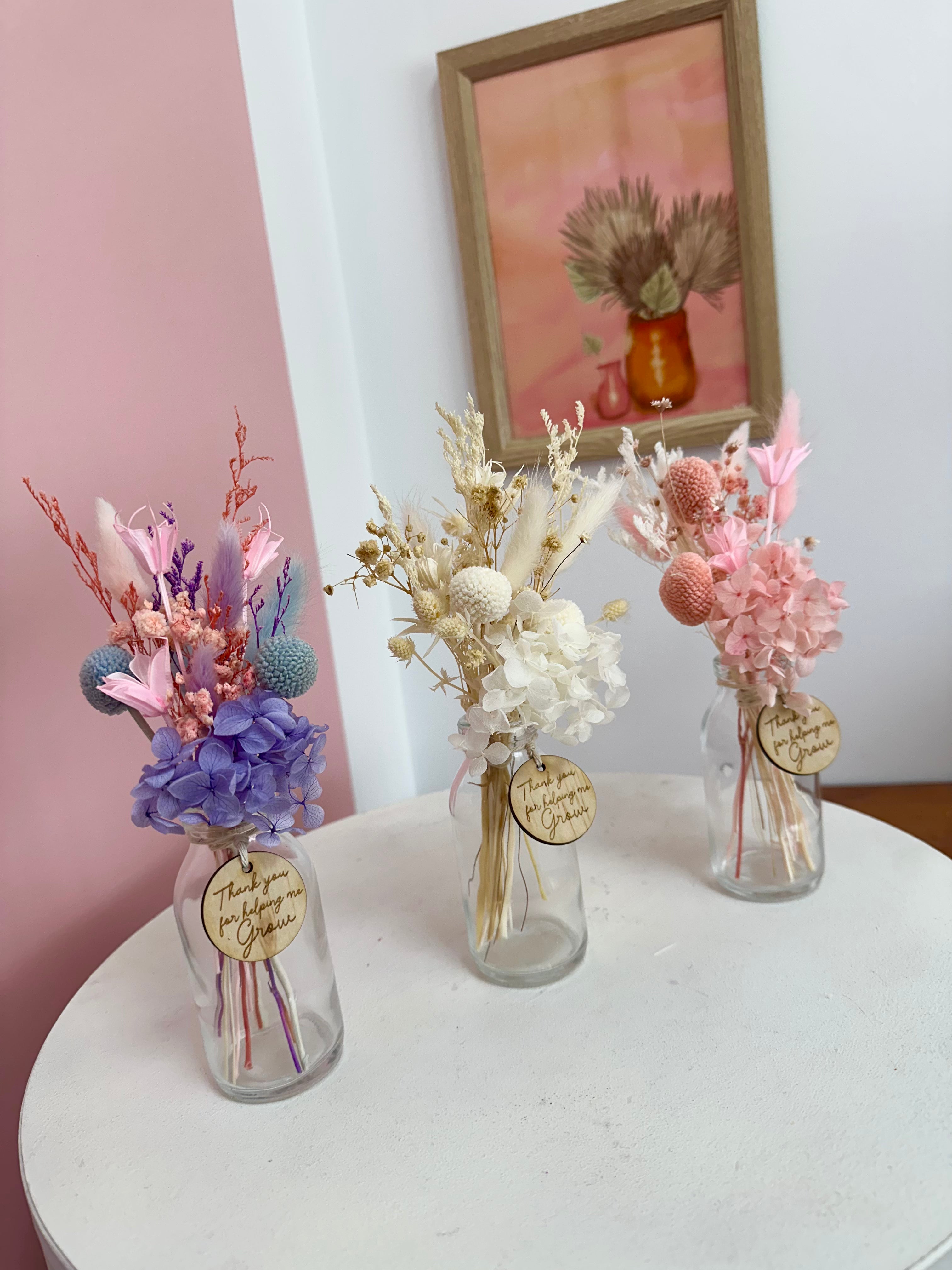 Three small glass vases with dried flowers on a white surface, with a framed picture in the background.