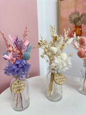Three glass vases with dried flowers and gold tags on a white surface against a pink wall.