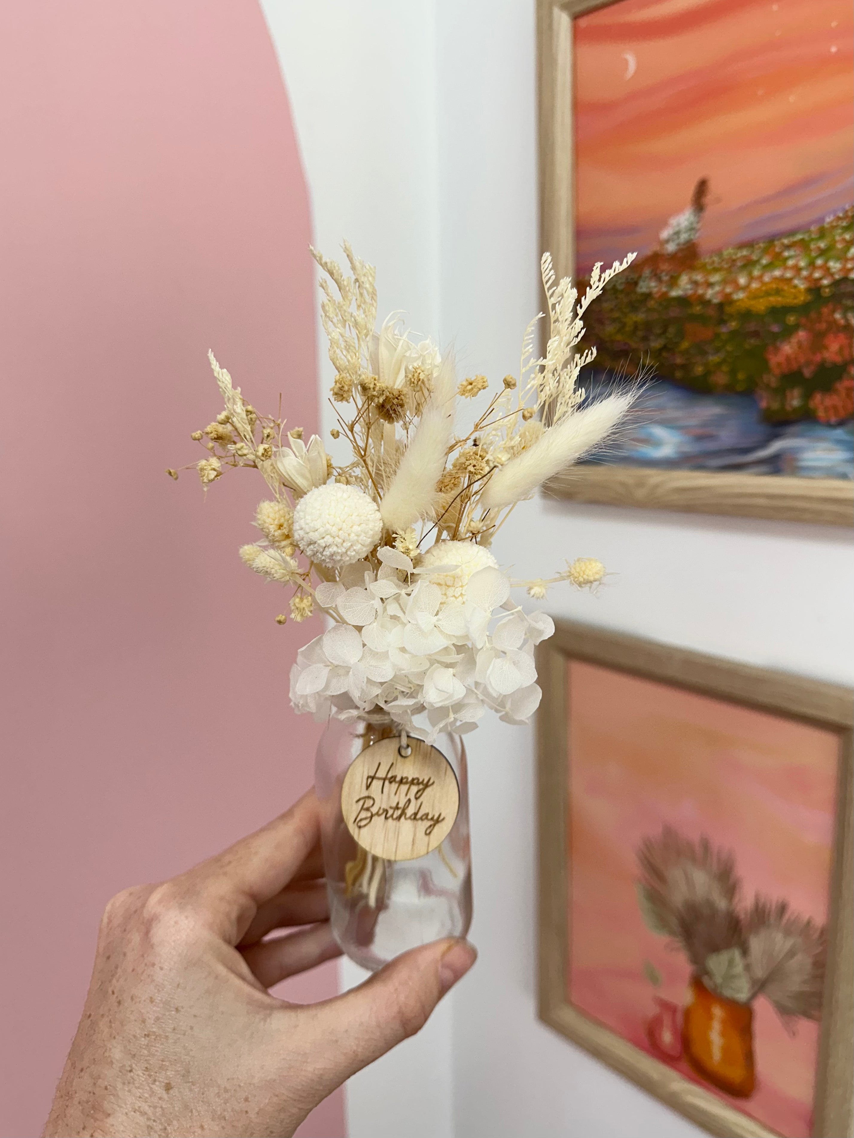 Hand holding a small glass vase with white flowers and a 'Happy Birthday' tag, against a wall with framed artwork.
