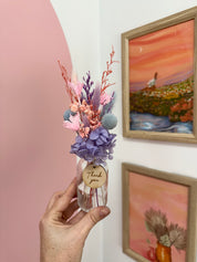 Hand holding a small bouquet of flowers with a 'thank you' tag in front of wall art.