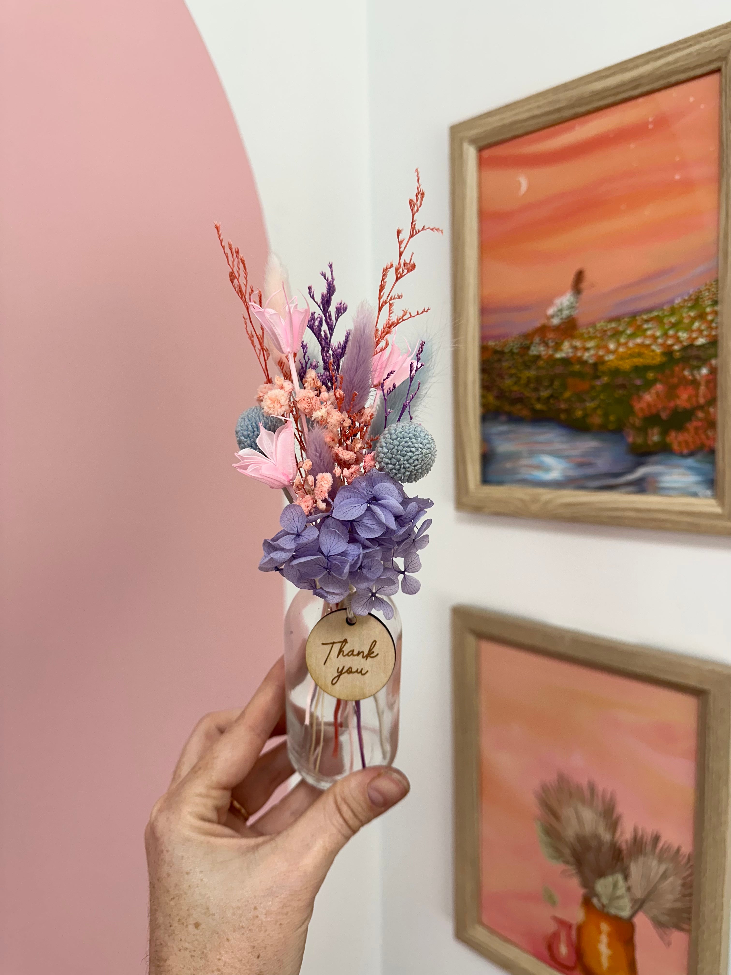 Hand holding a small bouquet of flowers with a 'thank you' tag in front of wall art.
